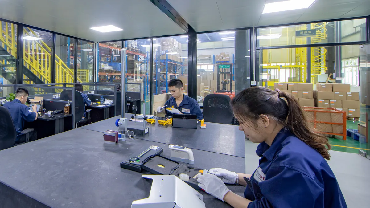Komaspec quality inspector in gloves measuring a sheet metal sample at an inspection bench, with gauges and a tablet