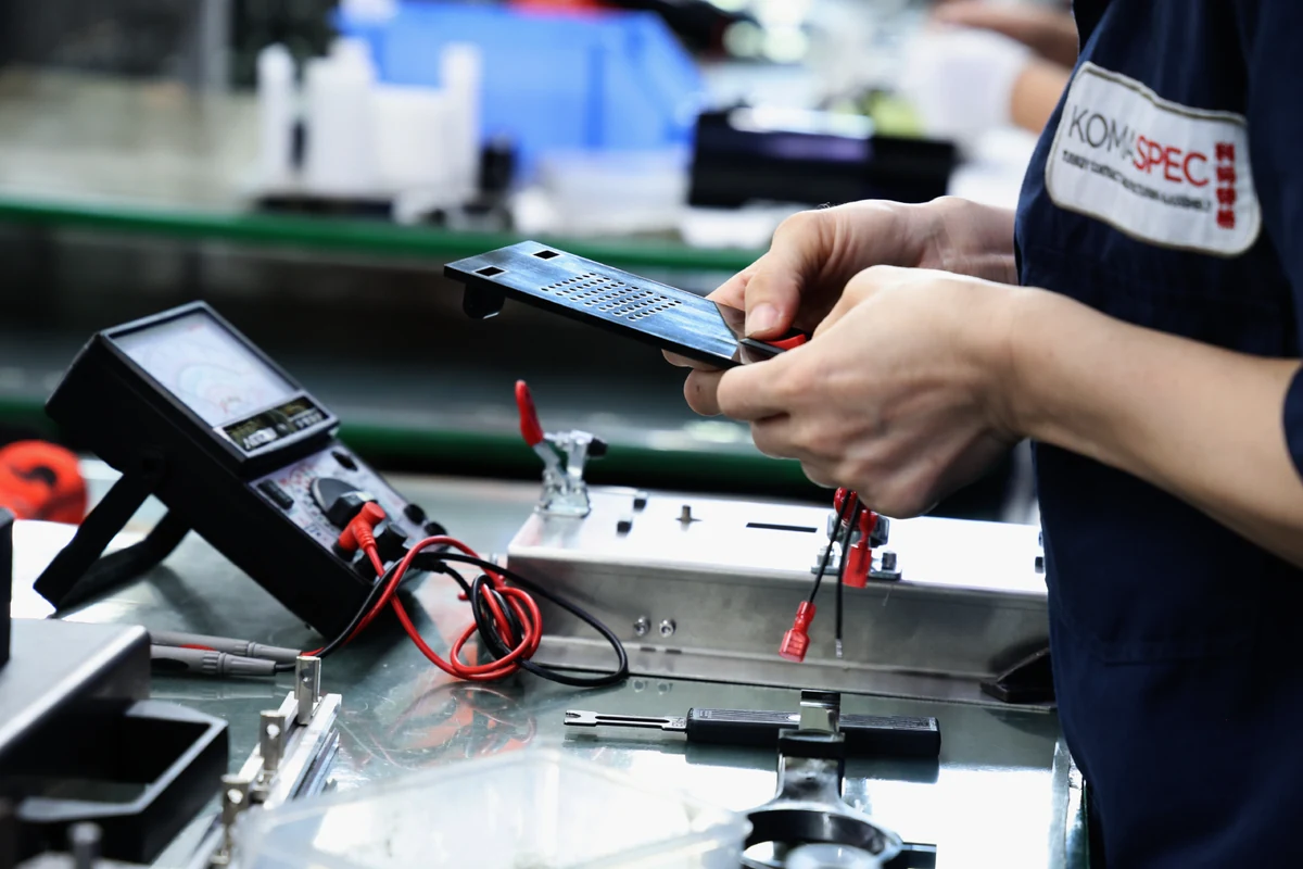 Technician testing a control panel and wiring with a digital multimeter at a Komaspec test bench