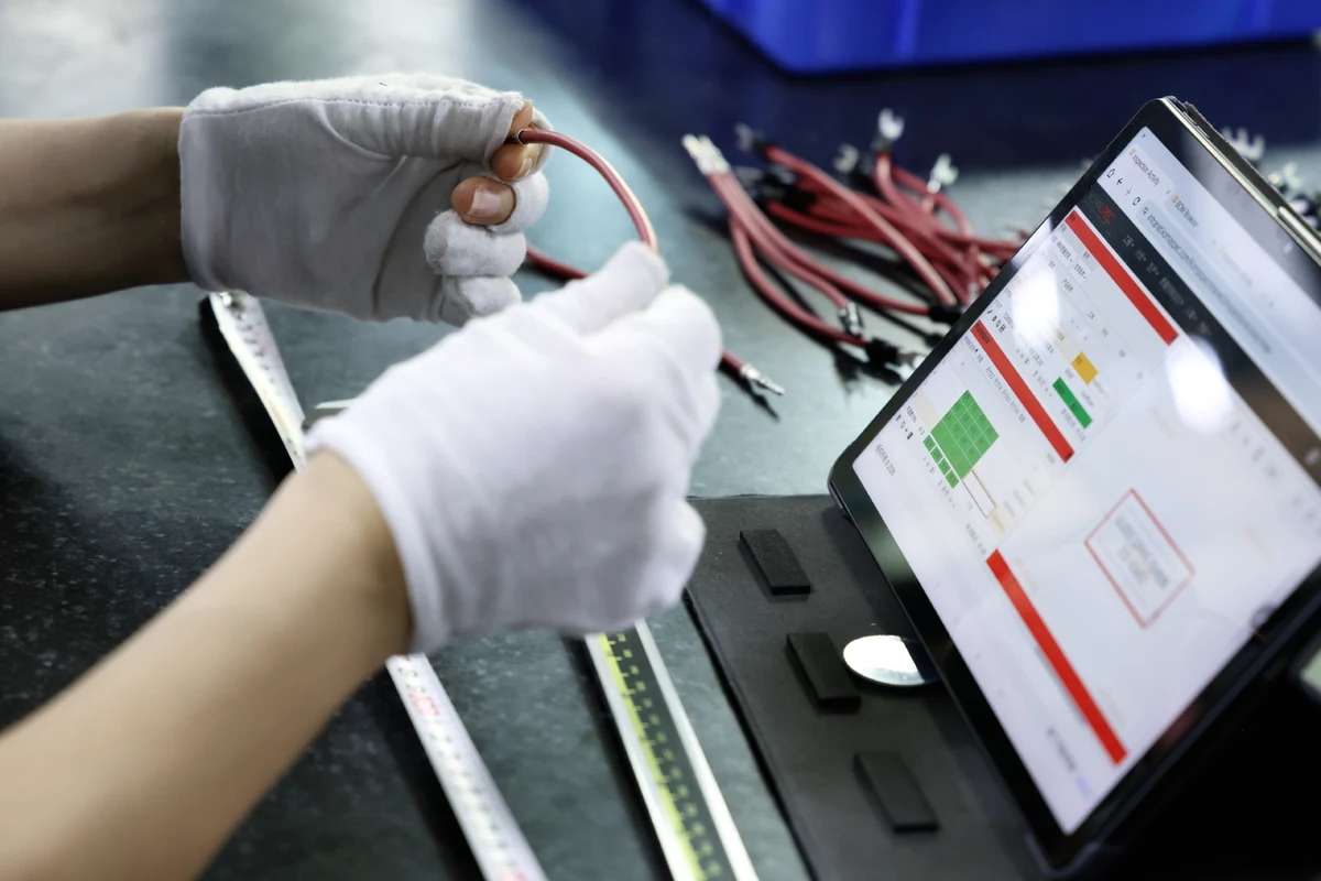 Gloved technician checking a red and black wire harness at an inspection station with a tablet showing pass and fail results