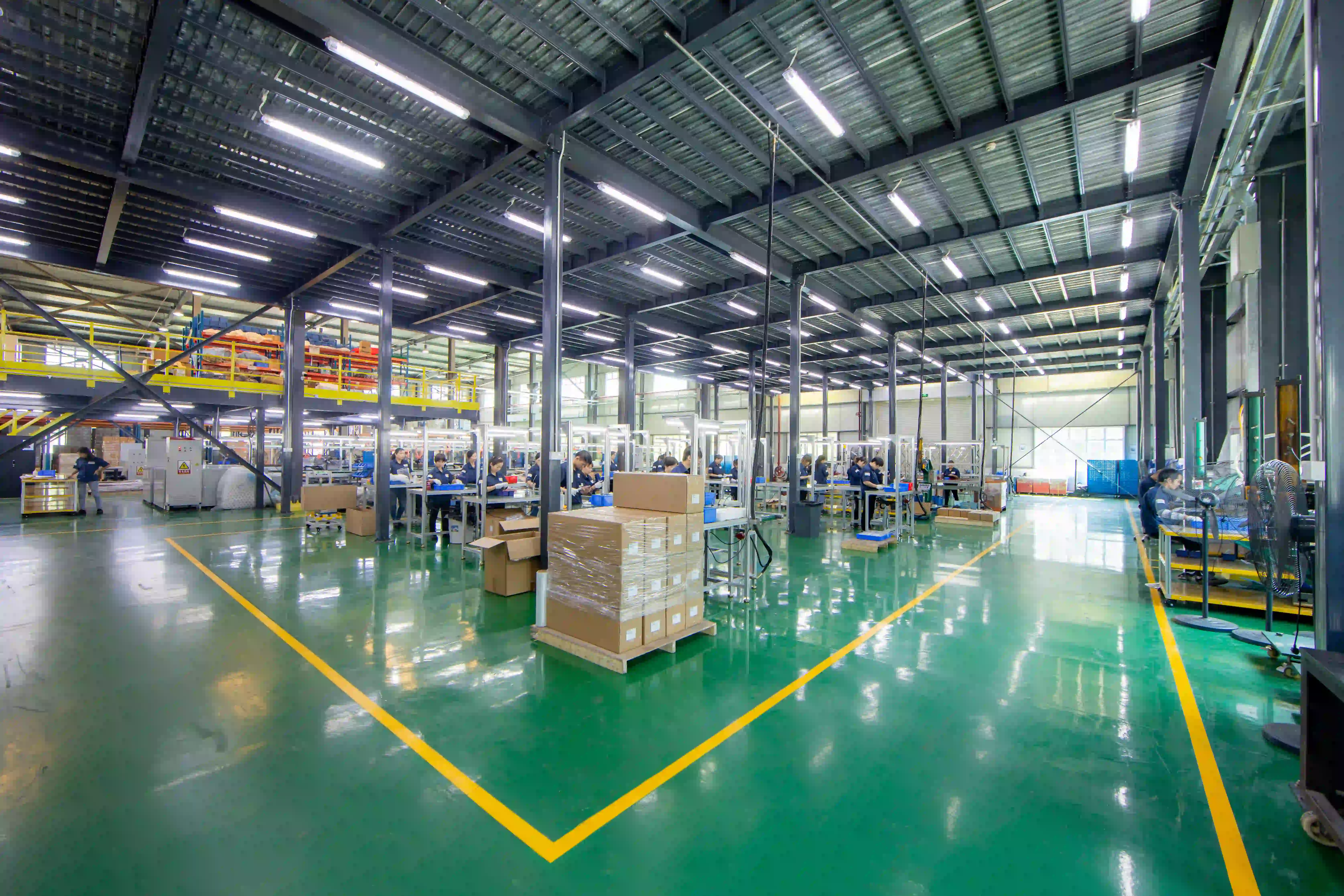 Stretch-wrapped pallet of cartons staged on a production floor, with assembly benches behind, showing packaging as a manufacturing BOM line
