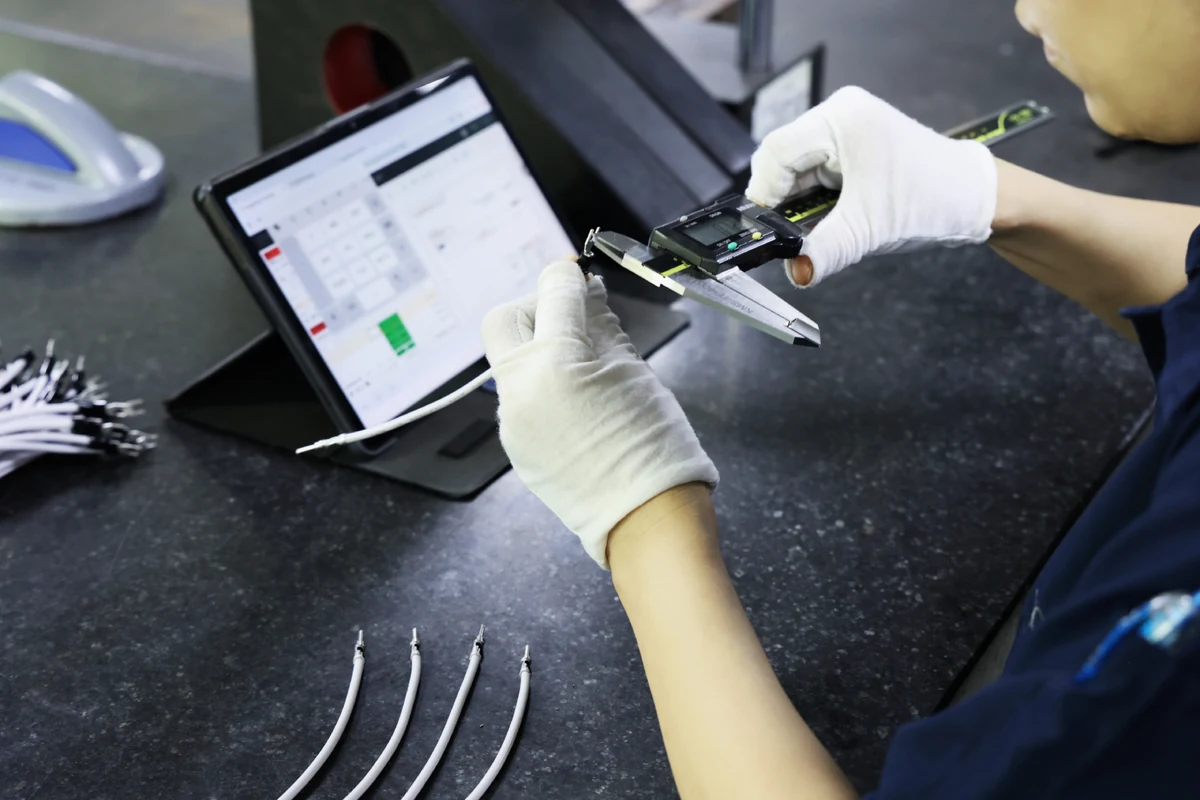Operator in ESD gloves measuring a populated module with digital calipers beside a tablet showing inspection results