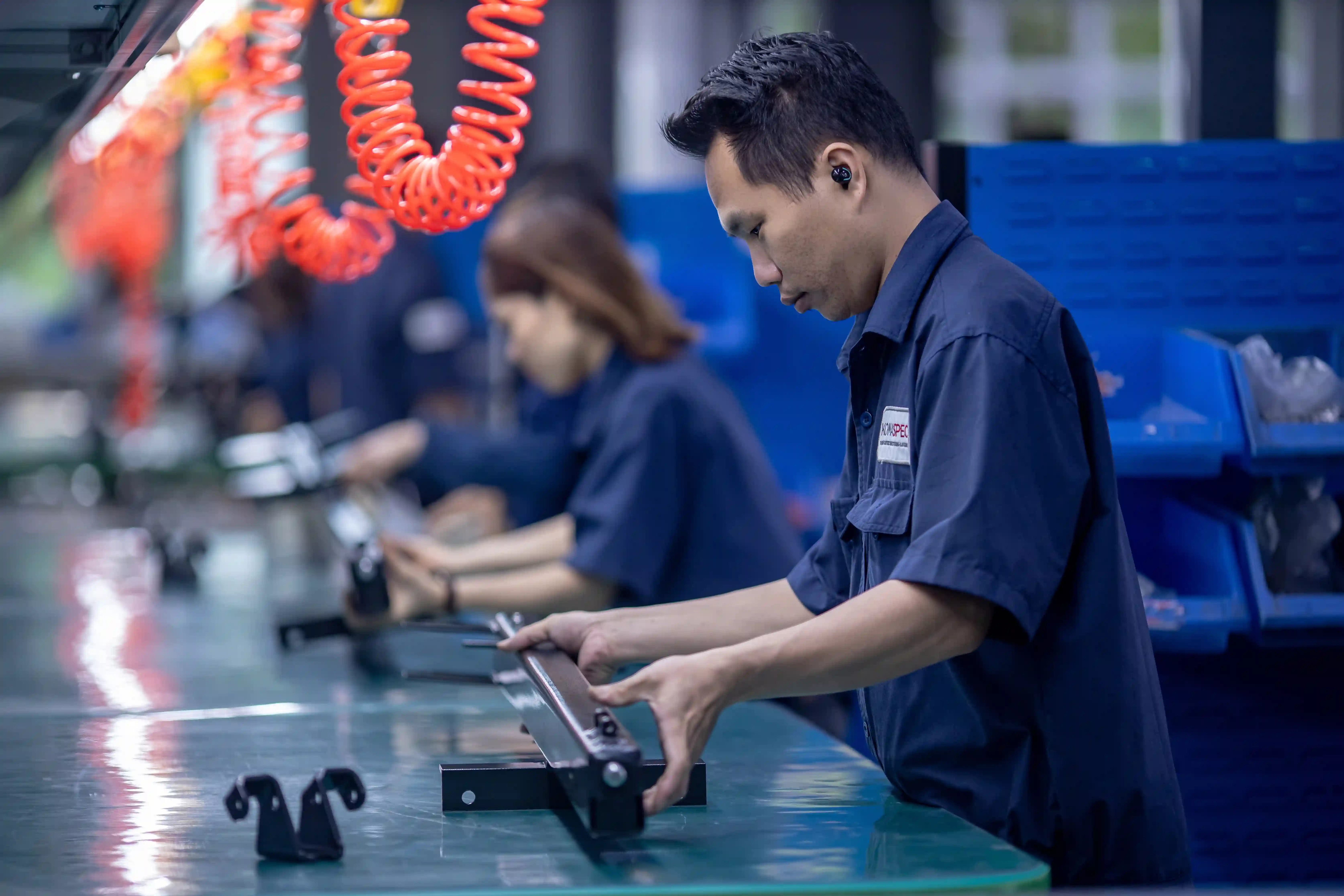 Assembly operator fitting a sheet metal part at a station, with the next brackets staged on the bench and component bins alongside