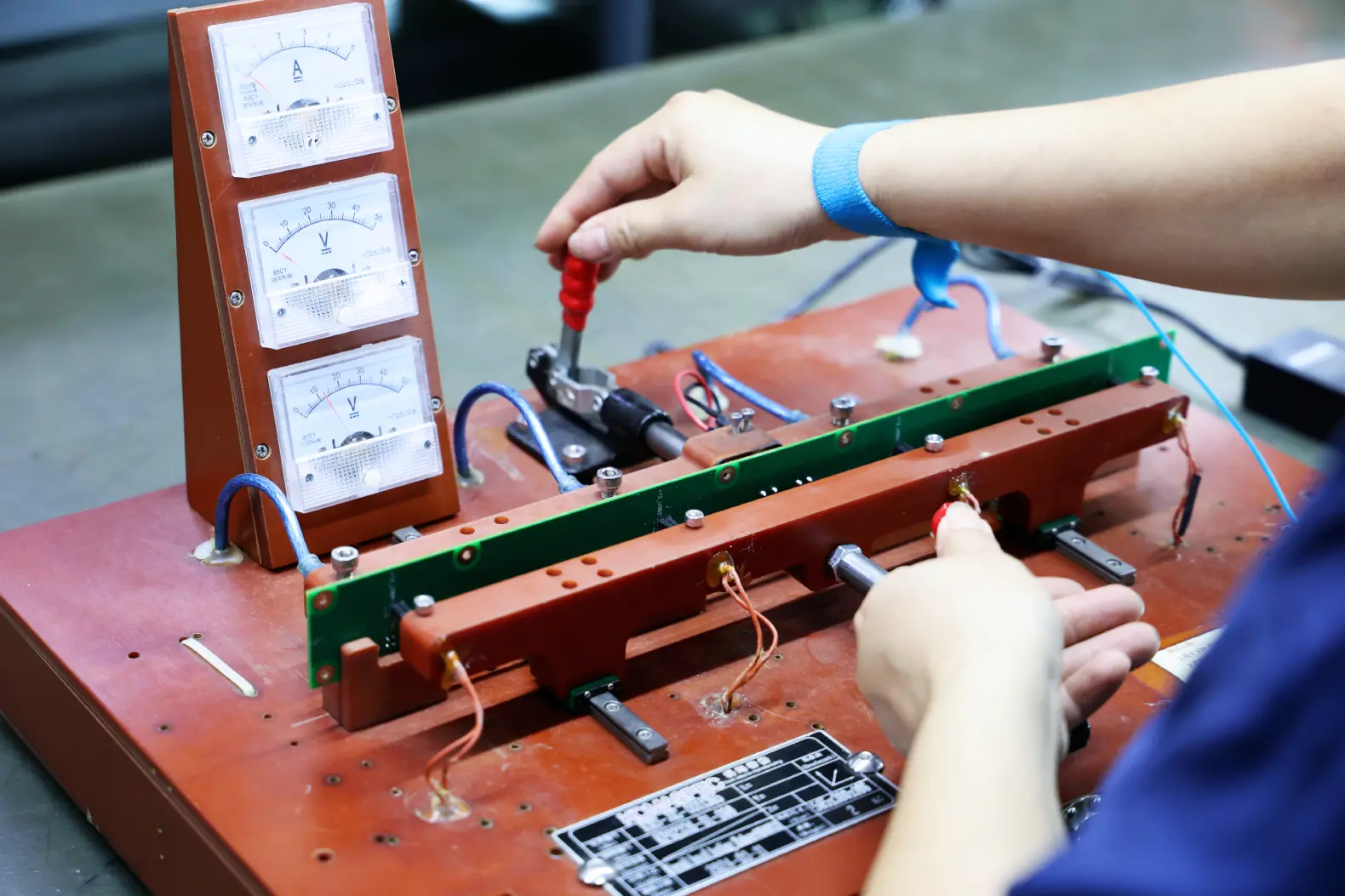 Operator running a functional test on a PCBA clamped into a custom jig with analogue current and voltage meters