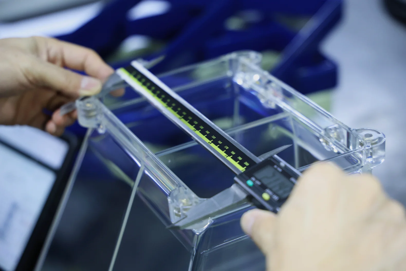 Technician measuring a clear injection-molded enclosure with digital calipers during dimensional inspection.
