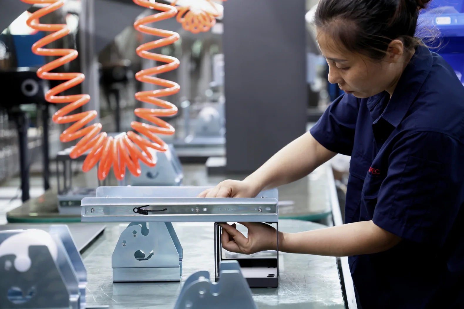Assembly worker fitting sheet metal parts into a fixture on a contract manufacturing line, with coiled air hoses overhead